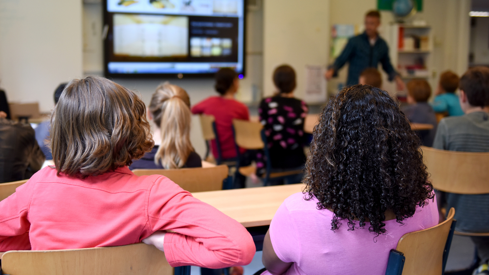 Two female students facing forward looking at teacher in front of classroom